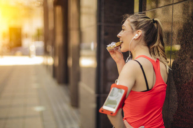 A woman facing away, wearing running attire, and eating a protein bar.