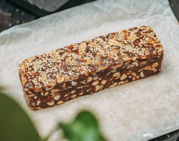 A brown protein bar, with light colored seeds and nuts in it, sitting on paper wrap