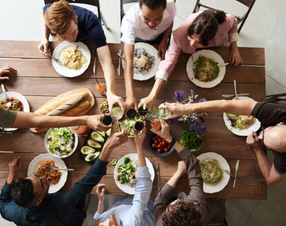 A picture taken from above, of 6 people sitting at a table with food on it, putting their glasses to the center of the table for a toast.