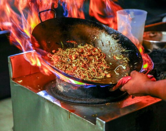 A picture of a person’s hand holding a pan with noodles and other food in it, with flames in the background.