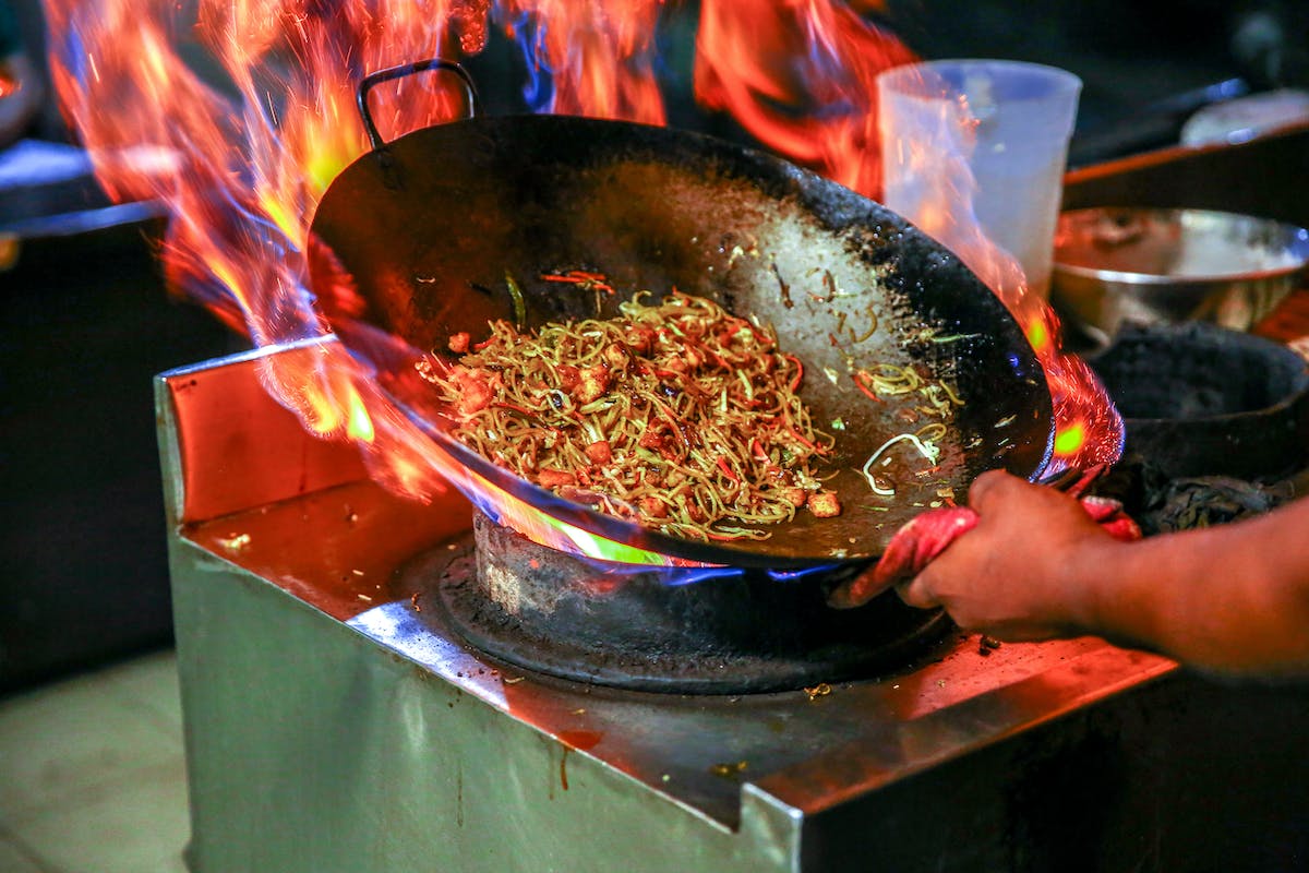 A picture of a person’s hand holding a pan with noodles and other food in it, with flames in the background.