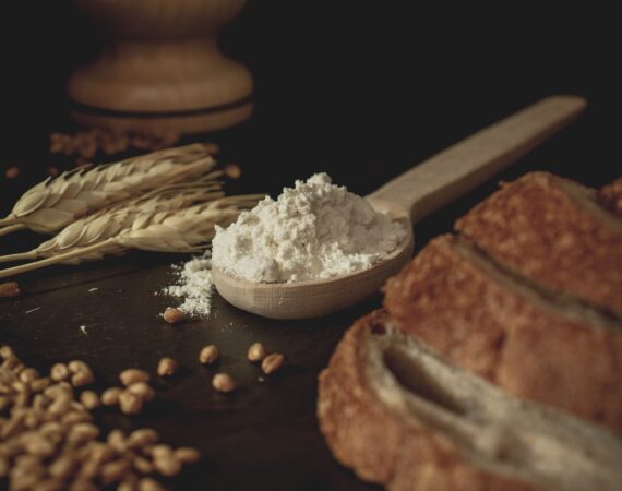 A picture of a spoon with flour in it, between a loaf of bread and grains of wheat.