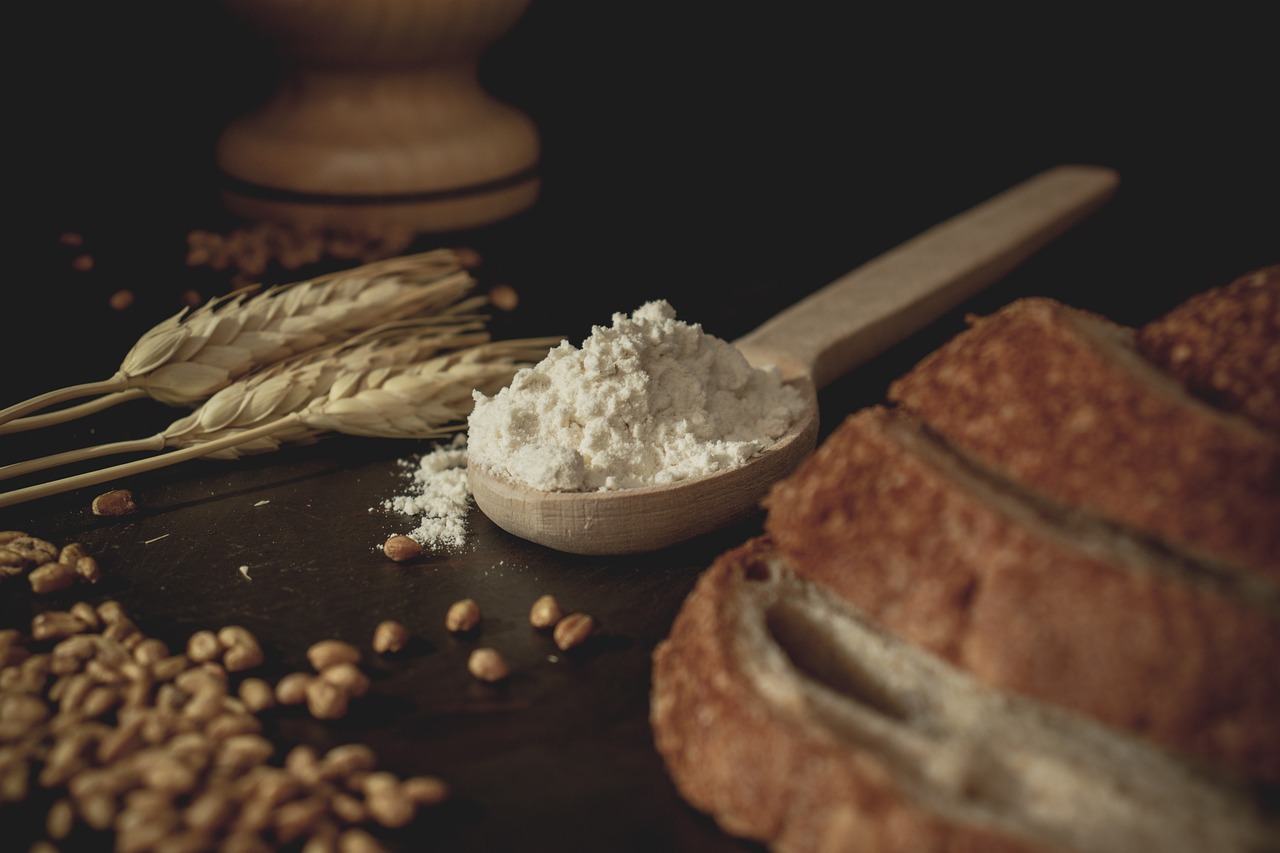 A picture of a spoon with flour in it, between a loaf of bread and grains of wheat.