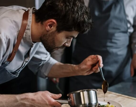 A picture of a chef watching another chef leaning over a dish and carefully pouring something into it from a spoon.